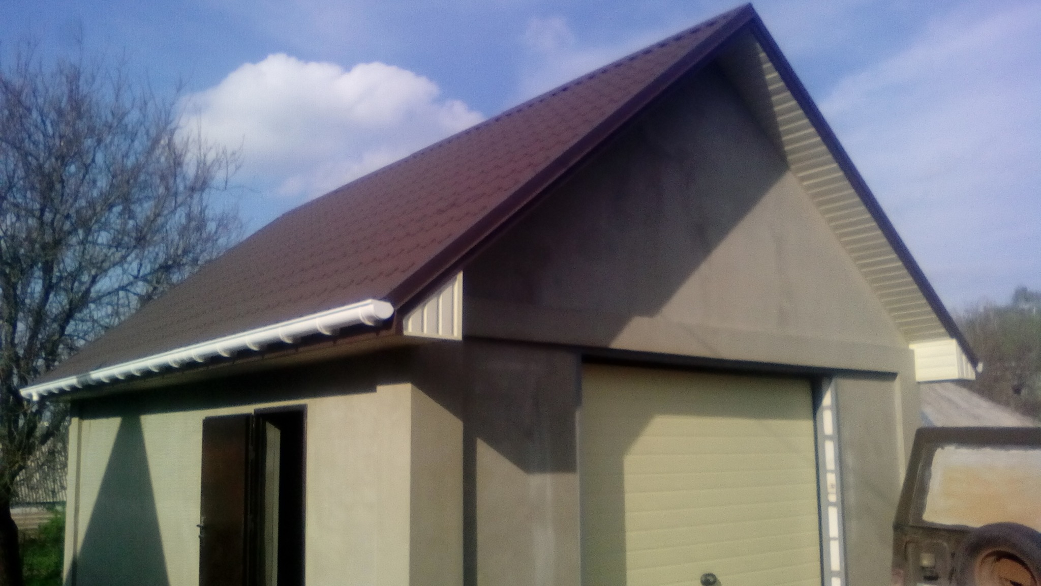 Garaż z brązowym dachem z blachodachówki, białymi rynnami i szarą elewacją. Widoczny fragment auta i drzewo bez liści w tle.