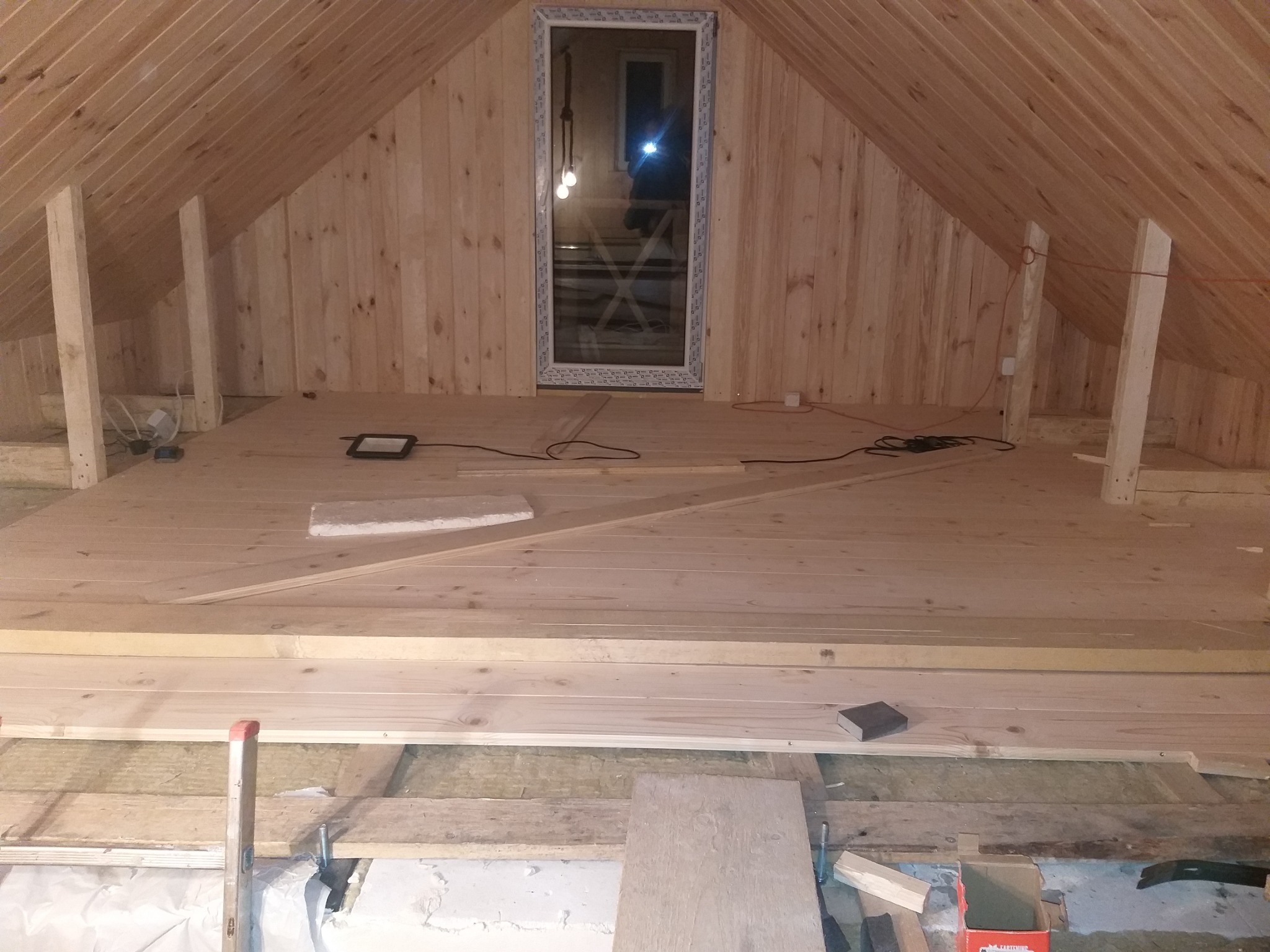 Poddasze w trakcie prac wykończeniowych: drewniane deski na podłodze i ścianach, okno w szczycie, widoczne materiały izolacyjne i narzędzia. Prace stolarskie.