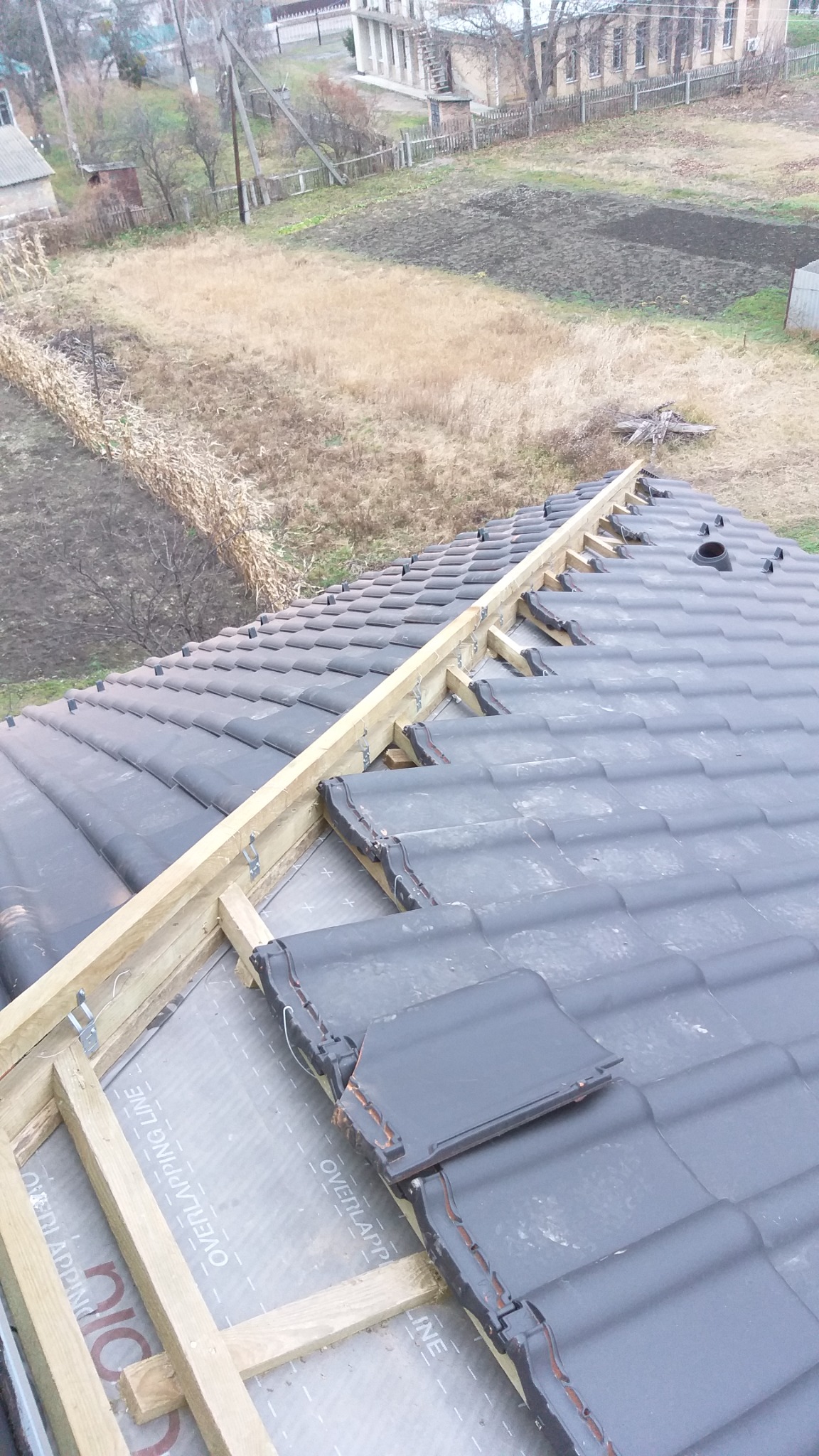 Montaż dachówki ceramicznej na drewnianej konstrukcji z folią w tle wiejskiego krajobrazu. Widok z góry na fragment dachu w trakcie prac dekarskich.