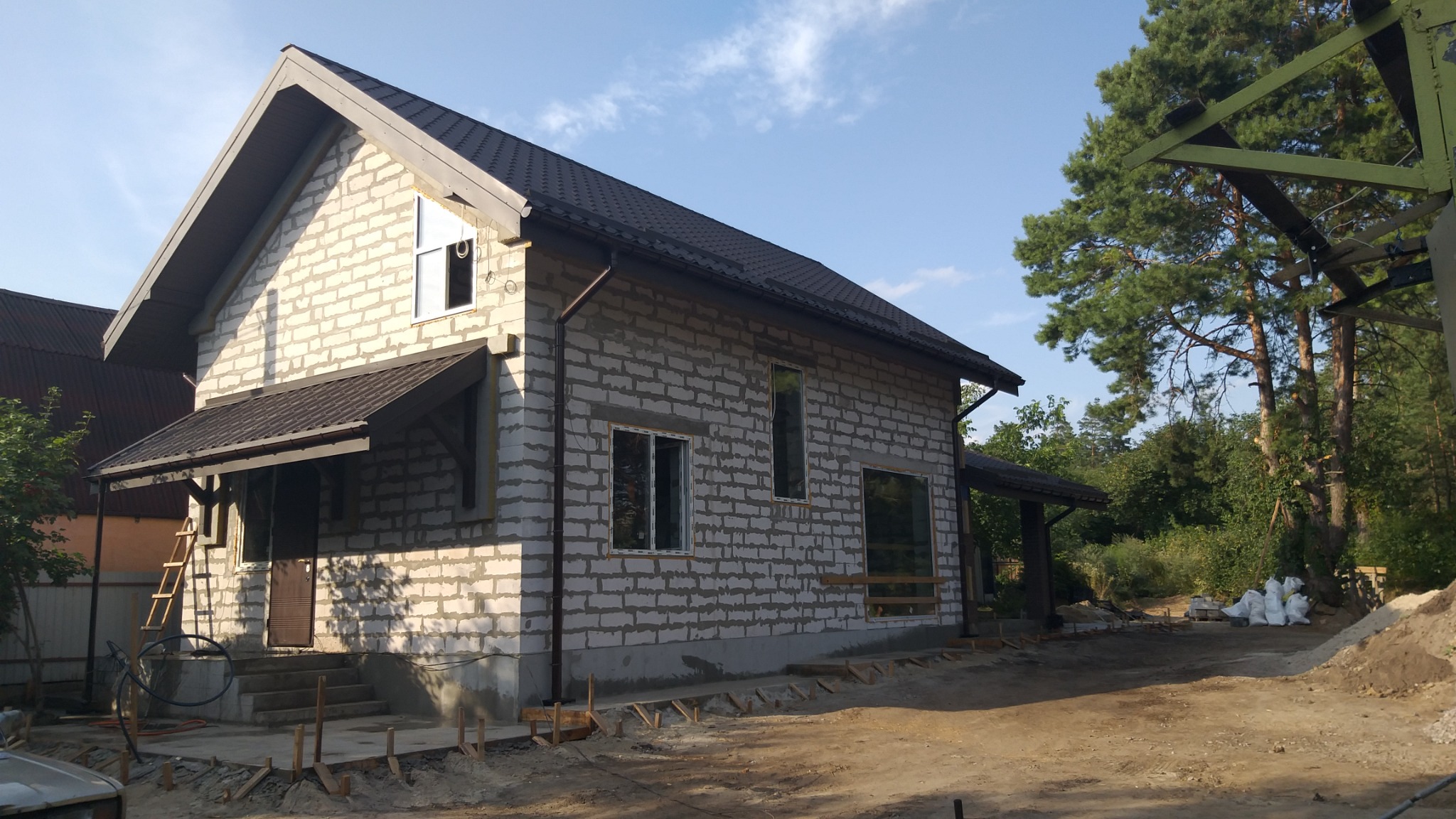 Dom murowany w trakcie budowy: ściany z betonu komórkowego, ciemna dachówka, widoczne okna i zadaszenie nad wejściem. Teren wokół w trakcie przygotowania.