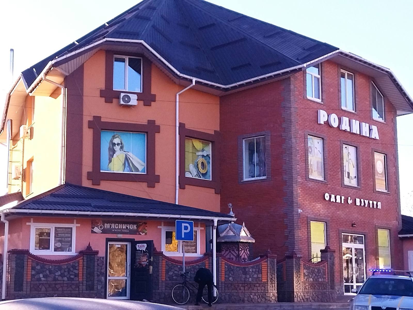 Dwupiętrowy budynek z ciemnym dachem, brązową cegłą i pomarańczową fasadą. Reklamy w oknach i szyldy sklepowe na parterze. Zaparkowany rower przed budynkiem.