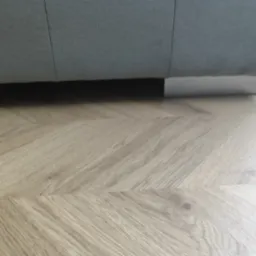 Ułożone panele podłogowe w jodełkę francuską. Jasne drewno podkreśla nowoczesny design wnętrza. Fragment sofy z szarej tkaniny z widocznymi srebrnymi nóżkami.