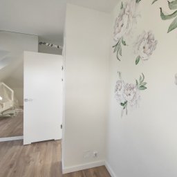 Home-Bud Grzegorz Musz - Jasne wnętrze poddasza z panelami, białą ścianą zdobioną kwiatowym motywem i otwartymi białymi drzwiami. Widoczne gniazdko elektryczne i listwa przypodłogowa.