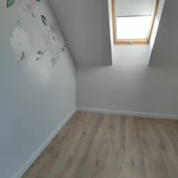 Jasne panele podłogowe w pokoju na poddaszu z oknem dachowym i ścianą ozdobioną delikatnymi, kwiatowymi naklejkami. Minimalistyczne wykończenie wnętrza.