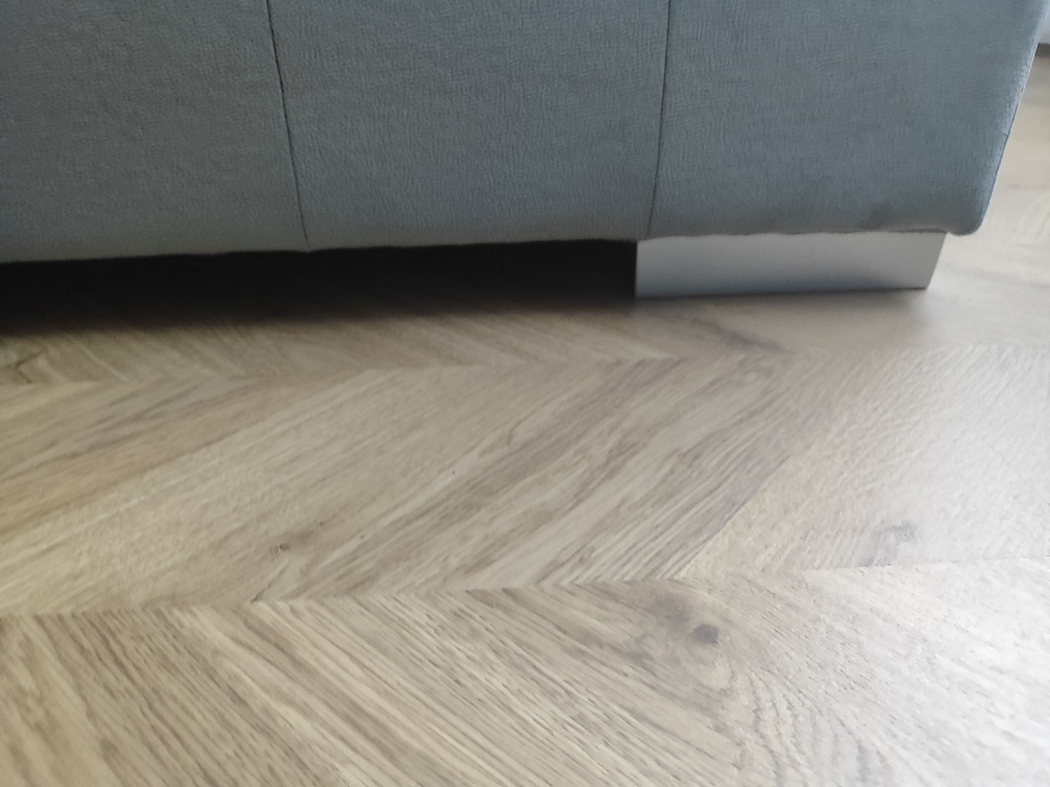 Ułożone panele podłogowe w jodełkę francuską. Jasne drewno podkreśla nowoczesny design wnętrza. Fragment sofy z szarej tkaniny z widocznymi srebrnymi nóżkami.