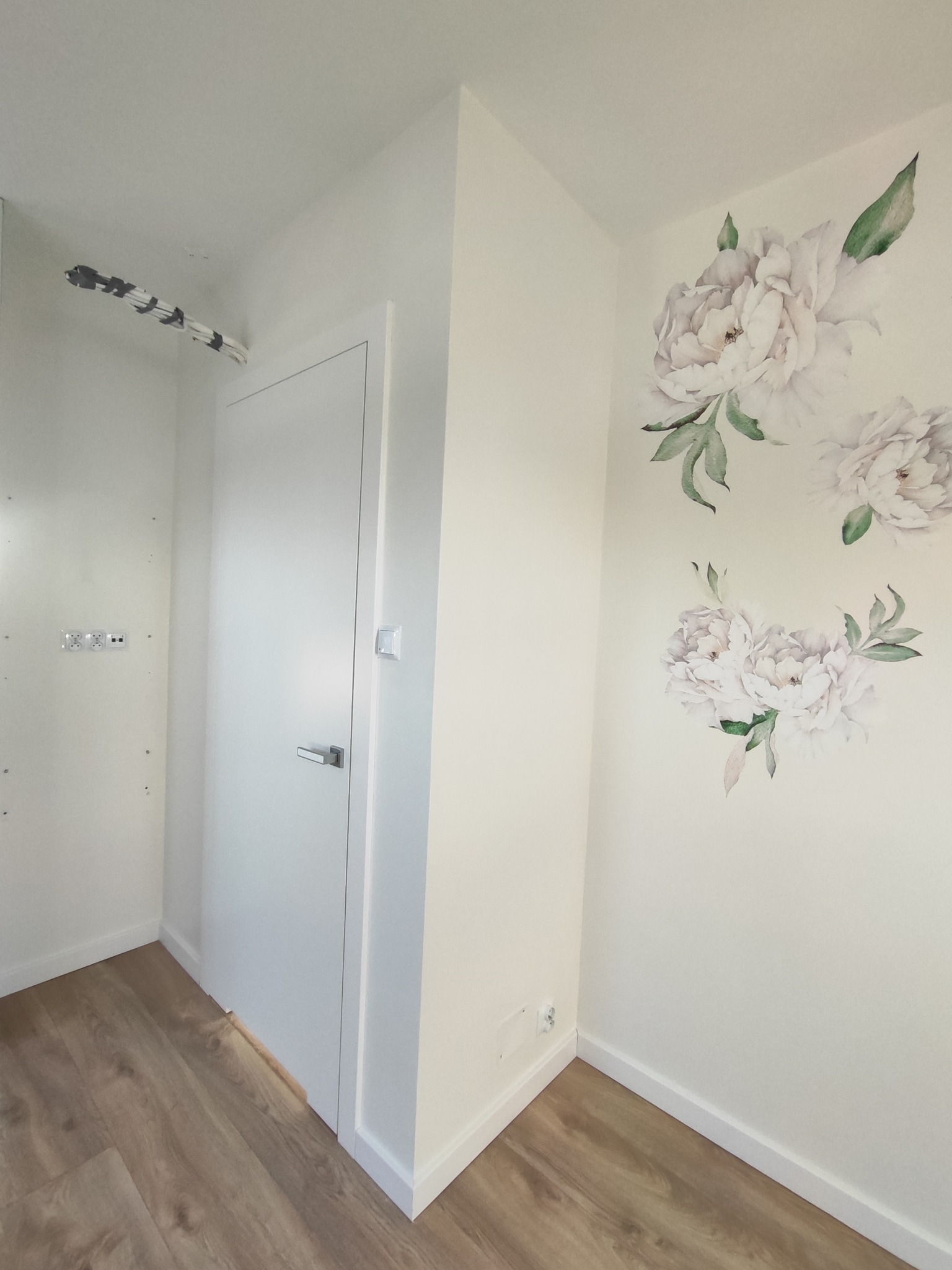 Białe drzwi wewnętrzne w korytarzu z panelami podłogowymi i dekoracyjnymi kwiatami na ścianie. Widoczne kable pod sufitem i gniazdka elektryczne.