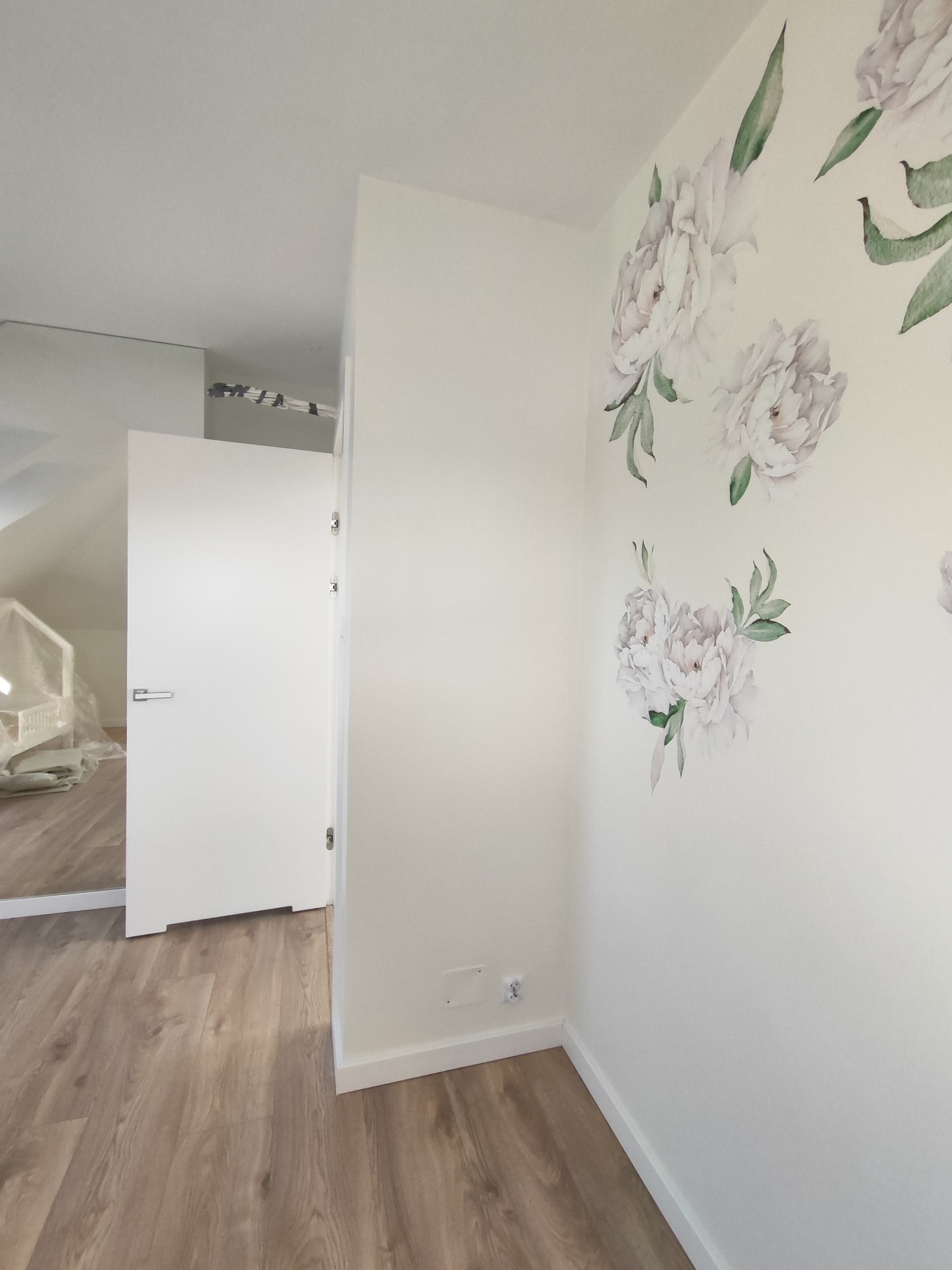 Jasne wnętrze poddasza z panelami, białą ścianą zdobioną kwiatowym motywem i otwartymi białymi drzwiami. Widoczne gniazdko elektryczne i listwa przypodłogowa.
