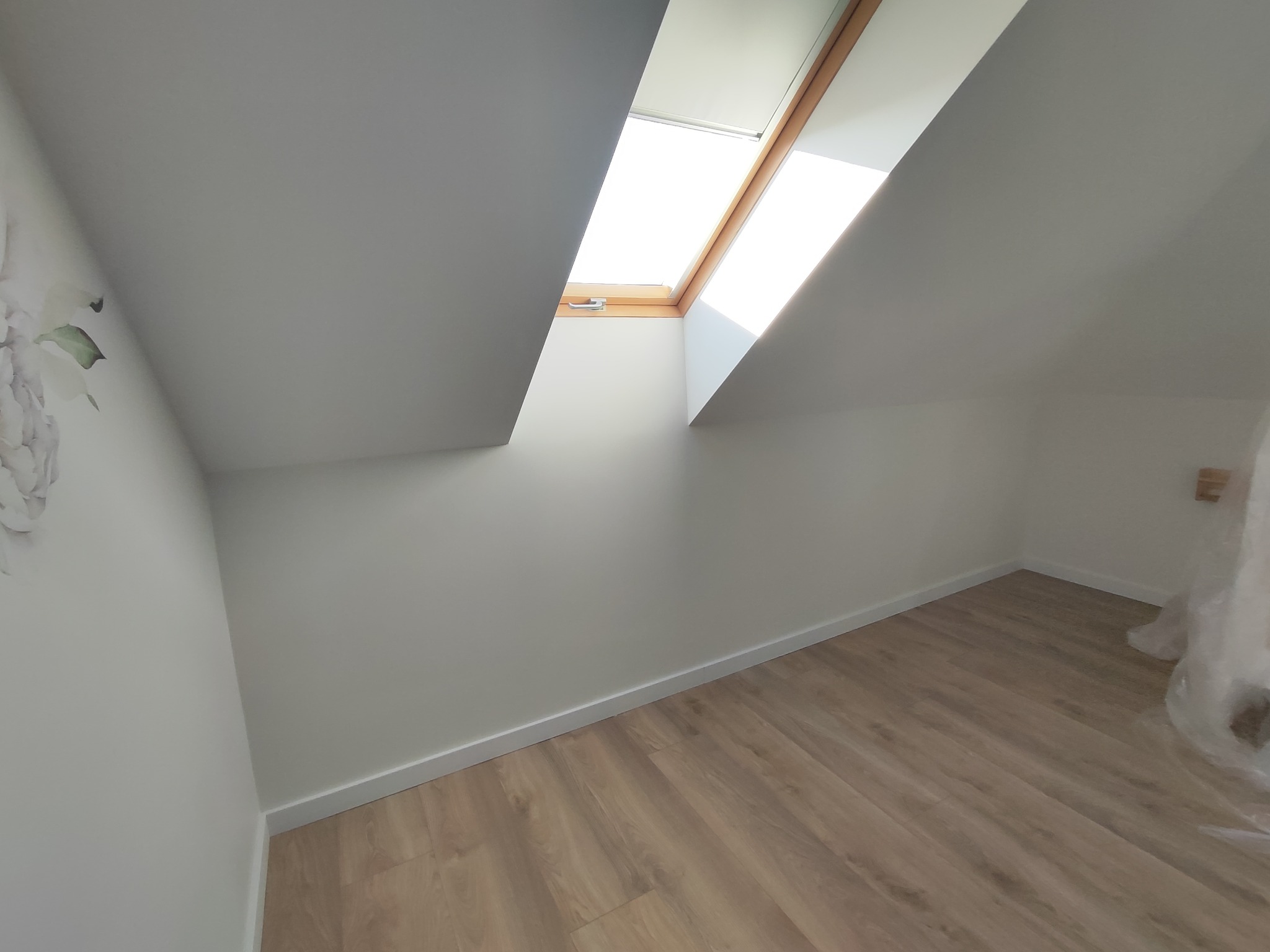 Poddasze po remoncie: białe ściany, drewniana podłoga i okno dachowe wpuszczające jasne światło. Na ścianie delikatny motyw kwiatowy.