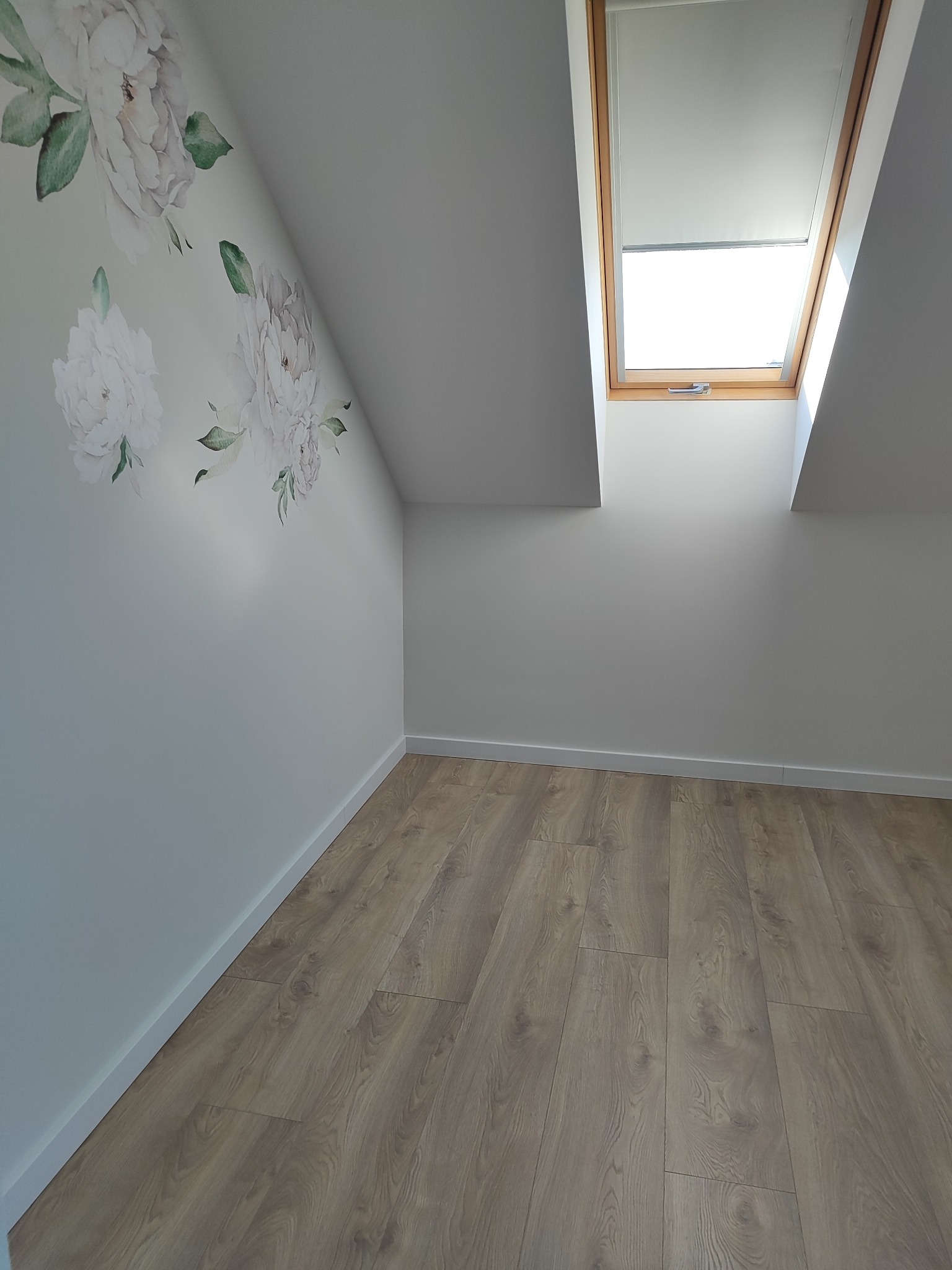 Jasne panele podłogowe w pokoju na poddaszu z oknem dachowym i ścianą ozdobioną delikatnymi, kwiatowymi naklejkami. Minimalistyczne wykończenie wnętrza.