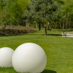 Ogrody D&T - Dwie białe, kuliste lampy ogrodowe na soczystej, zielonej trawie. W tle drzewa i ścieżka. Dekoracja ogrodu w słoneczny dzień.
