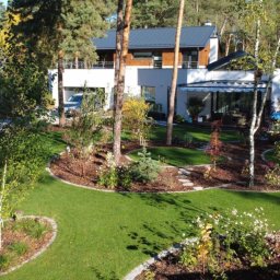 Ogrody D&T - Nowoczesny ogród z trawnikiem, rabatami i ścieżką z kamieni na tle białego domu z drewnianymi elementami. Widok z góry, słoneczny dzień.