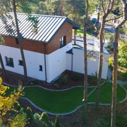 Ogrody D&T - Nowoczesny dom z ogrodem w Józefowie. Widok z góry na budynek z dwuspadowym dachem i starannie zaprojektowany trawnik z obrzeżami z kory i kamienia.