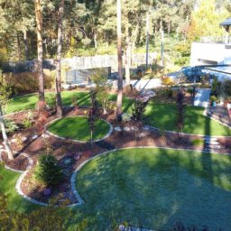 Ogrody D&T - Widok z góry na starannie zaprojektowany ogród z równo przystrzyżonym trawnikiem, rabatami z korą i kamieniami, drzewami oraz nowoczesnym domem w tle.