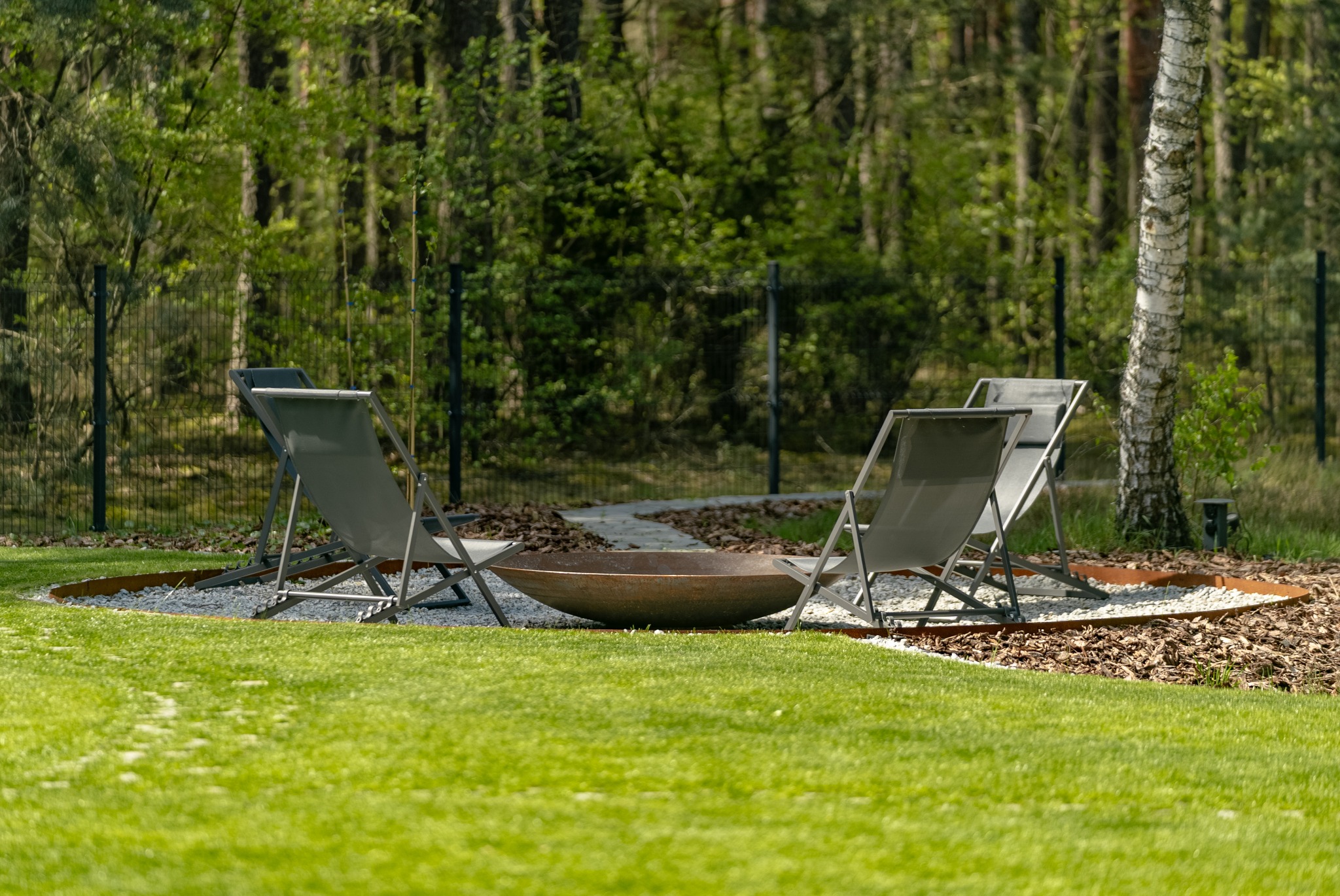 Ogród z paleniskiem otoczonym żwirem i korą, z dwoma leżakami. W tle las i metalowe ogrodzenie. Ujęcie z poziomu trawnika, w słoneczny dzień.