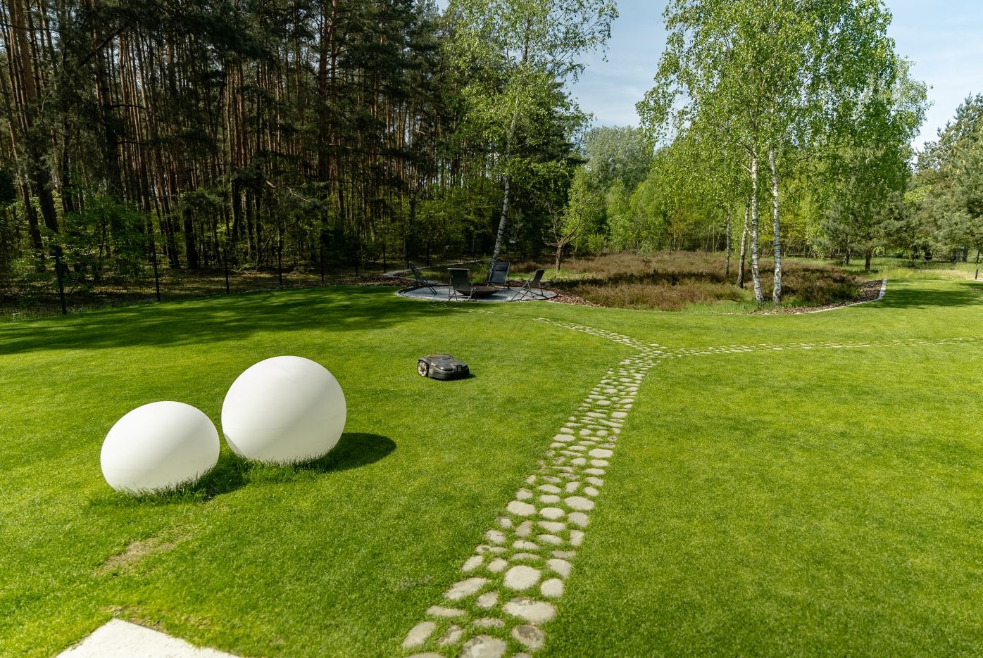 Zadbany trawnik z robotem koszącym, ścieżką z kamieni i dekoracyjnymi kulami. W tle drzewa i meble ogrodowe.