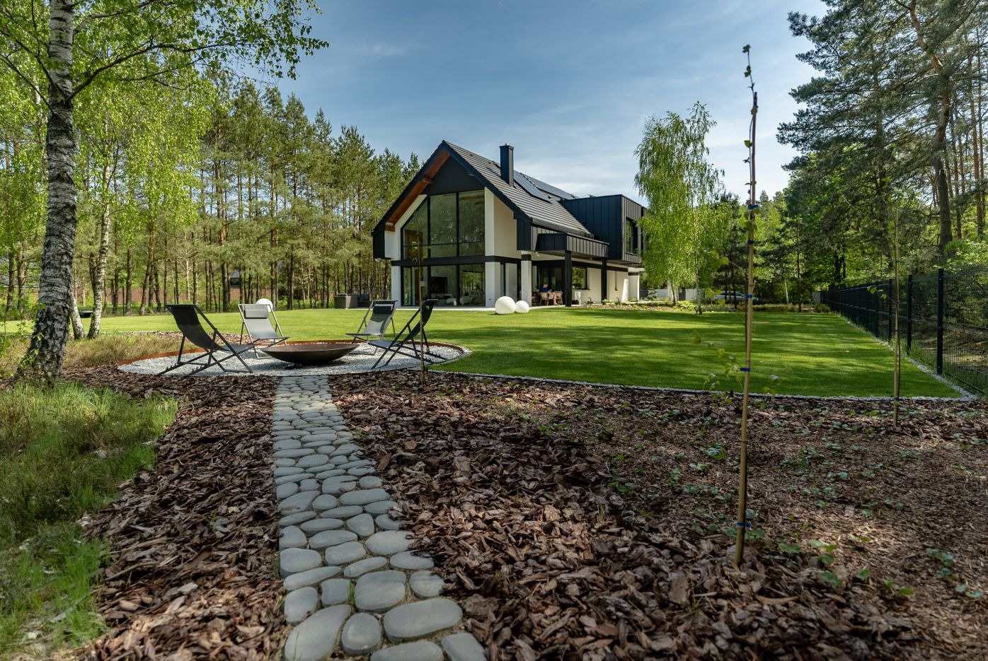 Nowoczesny dom z ogrodem: trawnik, ścieżka z kamienia prowadząca do paleniska otoczonego krzesłami. W tle las i minimalistyczna architektura budynku.