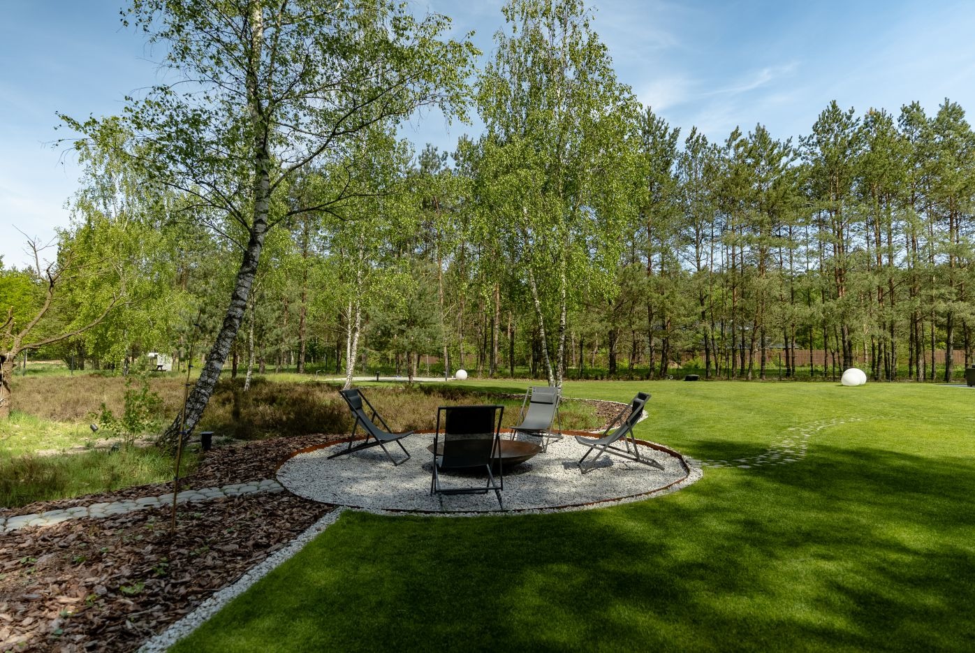 Ogród z okrągłym paleniskiem otoczonym białym żwirem i czterema krzesłami. W tle trawnik, drzewa i ścieżka z kory.