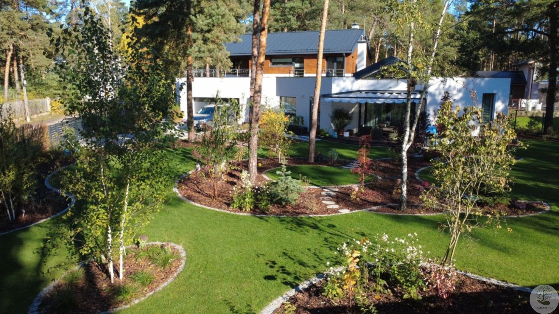 Nowoczesny ogród z trawnikiem, rabatami i ścieżką z kamieni na tle białego domu z drewnianymi elementami. Widok z góry, słoneczny dzień.