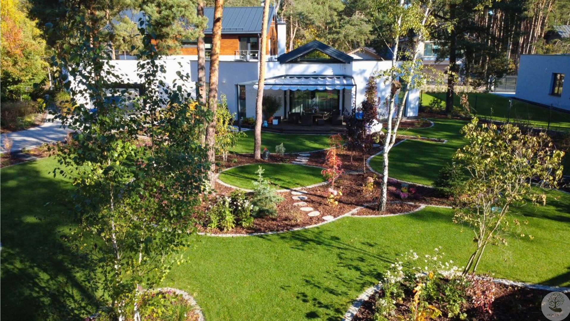 Zadbany ogród z równo przystrzyżonym trawnikiem, ozdobnymi rabatami i ścieżką z kamieni prowadzącą przez korę. W tle nowoczesny dom z tarasem.