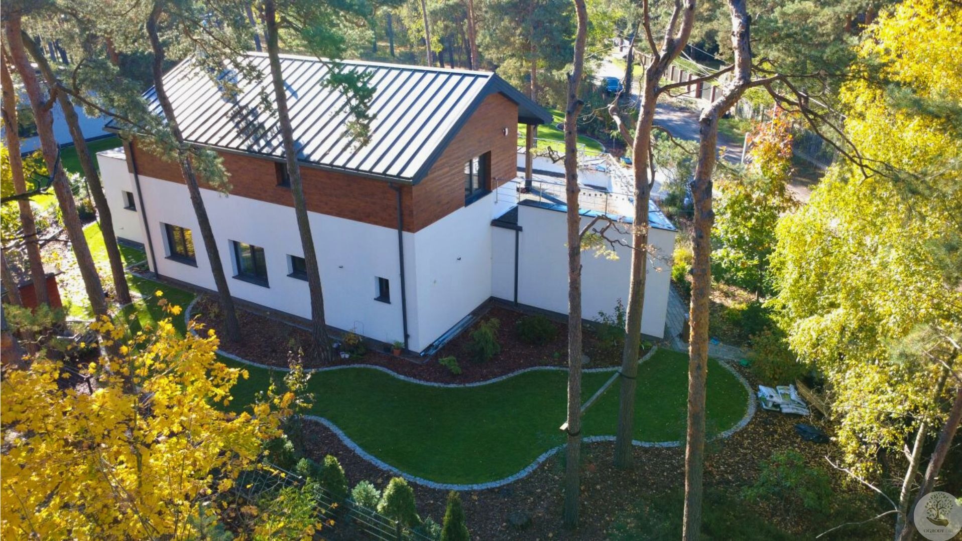 Nowoczesny dom z ogrodem w Józefowie. Widok z góry na budynek z dwuspadowym dachem i starannie zaprojektowany trawnik z obrzeżami z kory i kamienia.