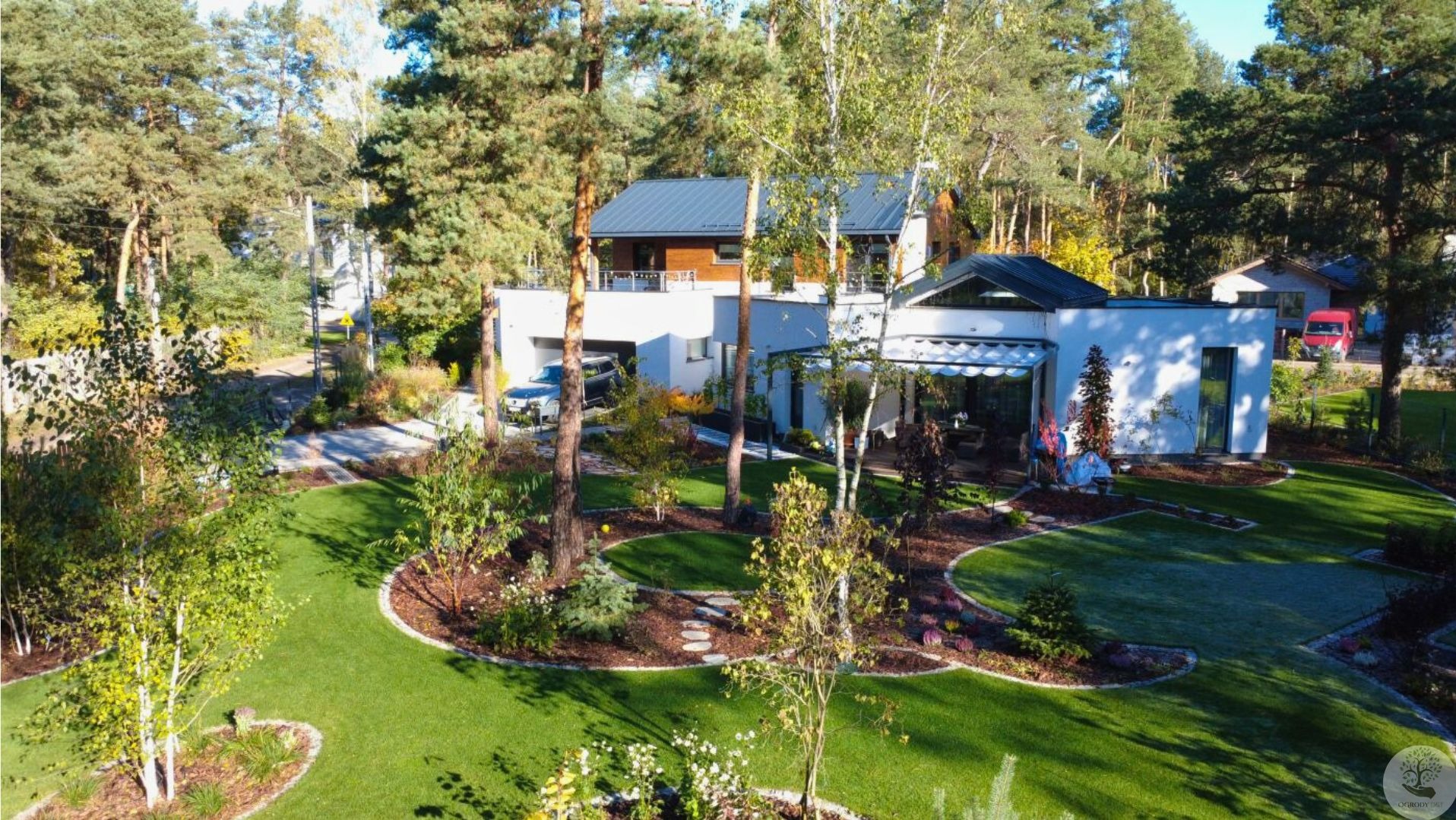 Rozległy, zadbany ogród z nowoczesnym domem w tle. Widoczny trawnik, drzewa, krzewy i ścieżki. Całość utrzymana w harmonijnej kolorystyce z elementami architektury krajobrazu.