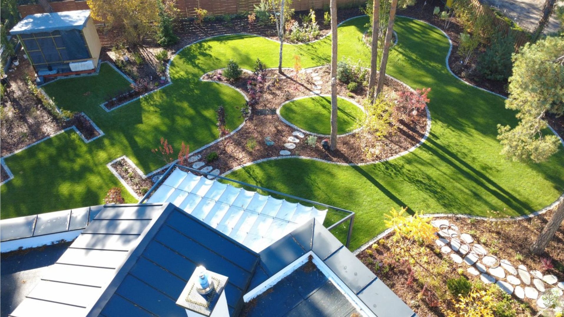 Widok z góry na starannie zaprojektowany ogród z trawnikiem, rabatami i ścieżkami z kamienia, otoczony drzewami i krzewami. W tle widoczny fragment dachu budynku.