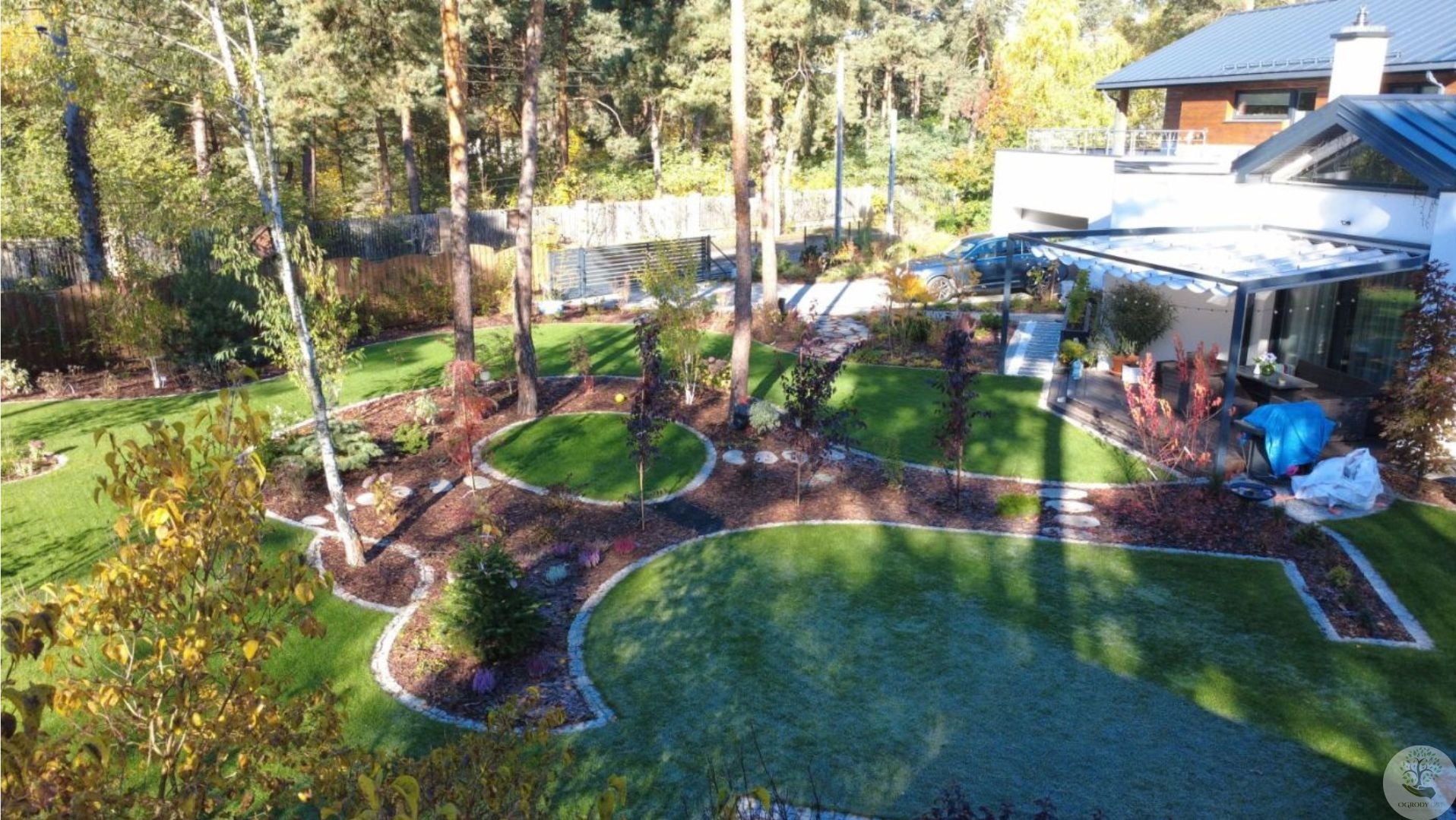 Widok z góry na starannie zaprojektowany ogród z równo przystrzyżonym trawnikiem, rabatami z korą i kamieniami, drzewami oraz nowoczesnym domem w tle.