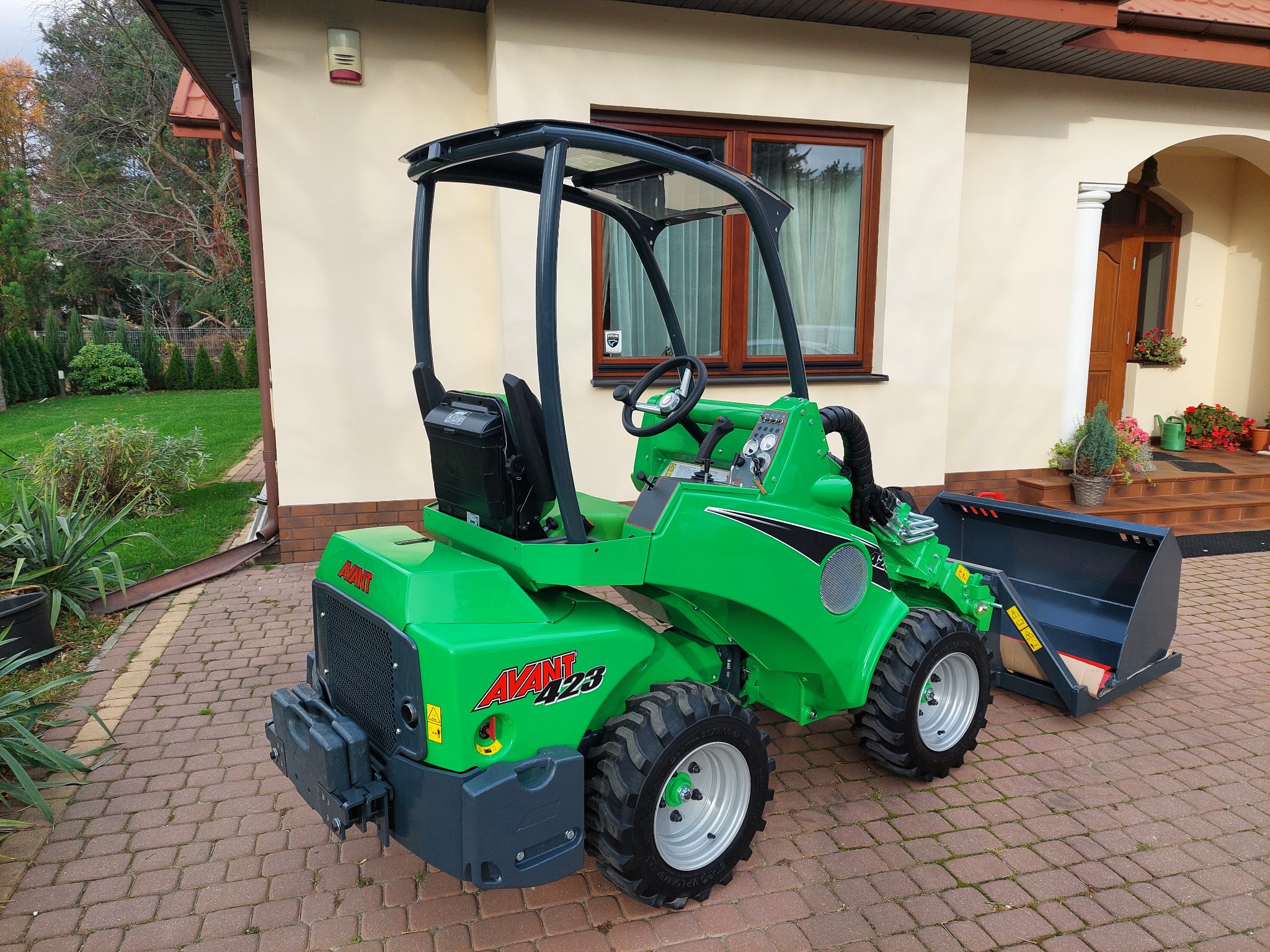 Zielona miniładowarka Avant 423 na kostce brukowej przed domem z beżową elewacją. Widoczny fragment ogrodu i okno z zasłonami.