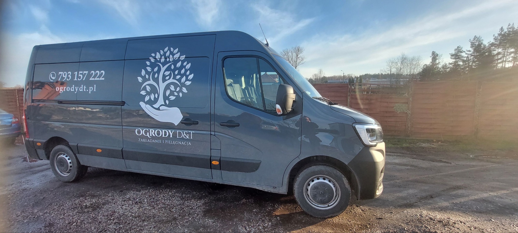Szary samochód dostawczy z logo firmy ogrodniczej 'Ogrody D&T' i numerem telefonu, zaparkowany na żwirowym podłożu, z drewnianym ogrodzeniem w tle.
