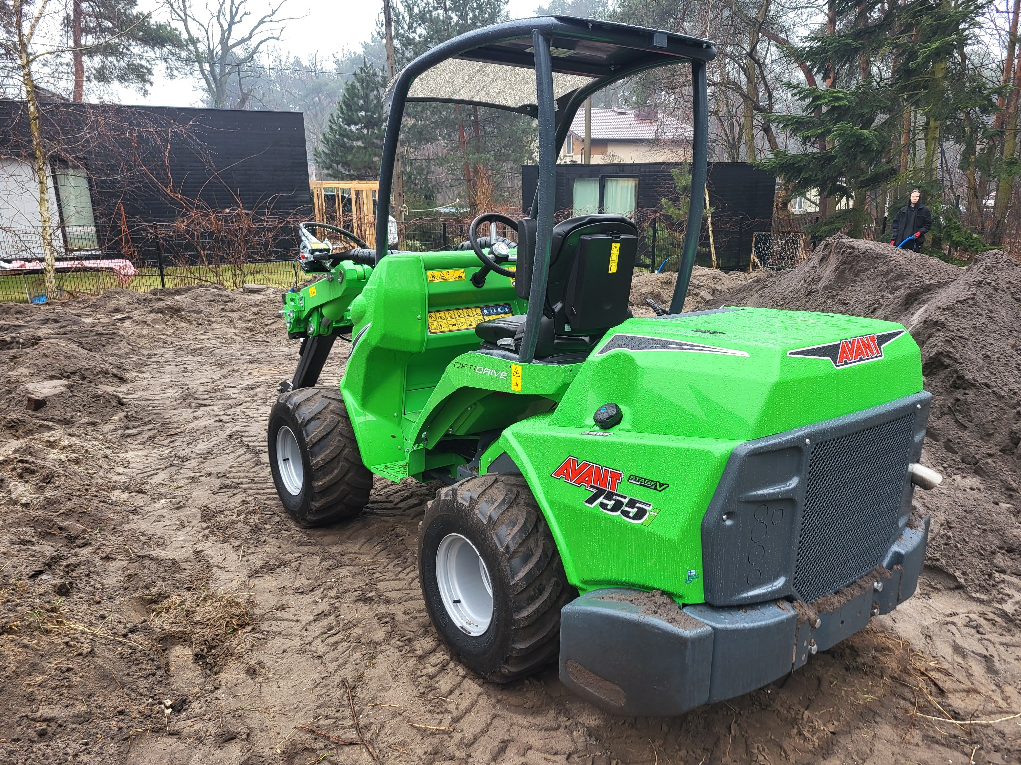 Zielona miniładowarka Avant 755 na placu budowy, z widocznymi śladami opon na błotnistym podłożu. W tle domy i drzewa, a w oddali osoba z niebieskim wężem.