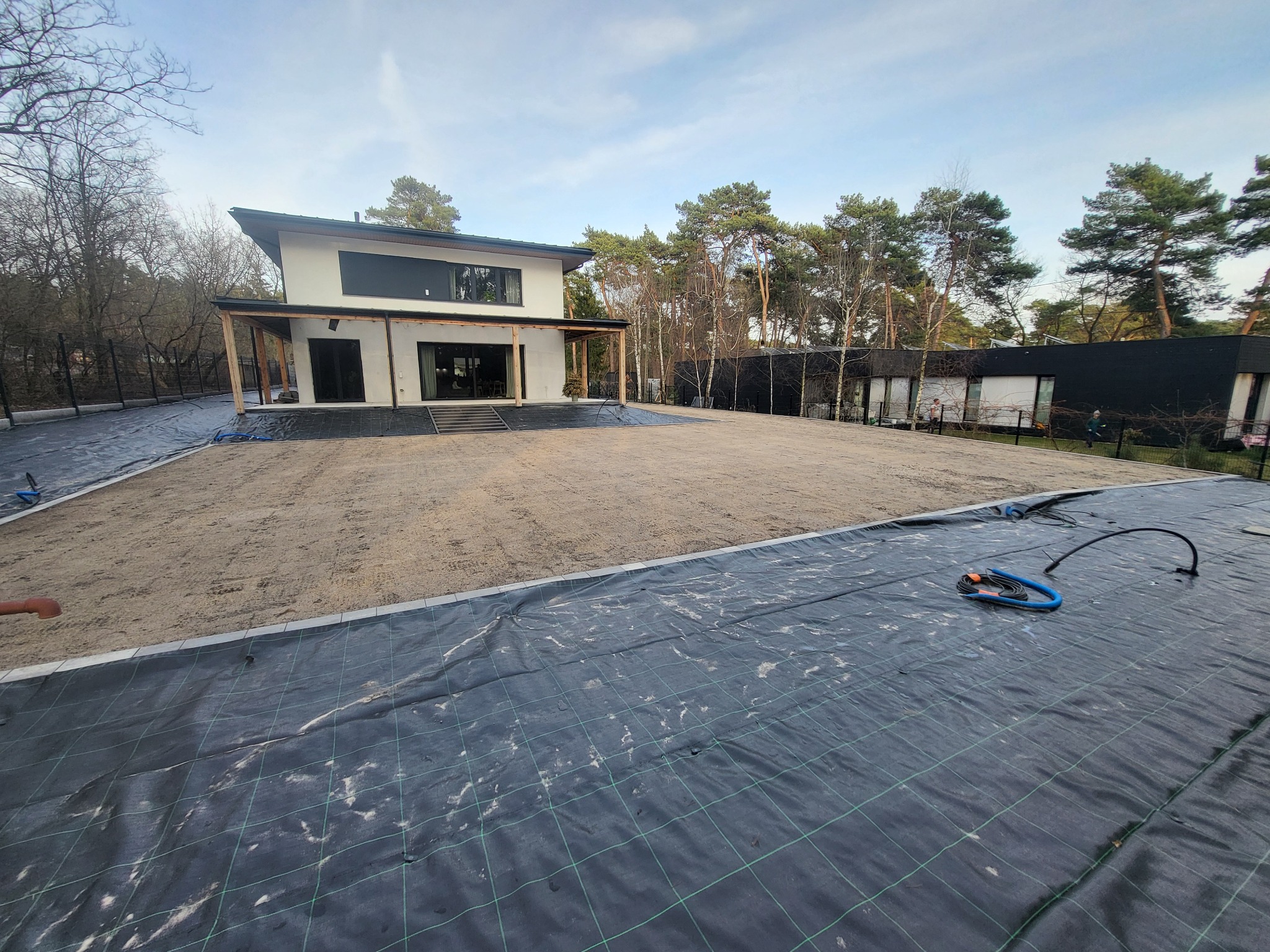 Przygotowanie terenu pod trawnik: rozłożona agrotkanina na piasku, obrzeża trawnikowe, nowoczesny dom w tle, drzewa. Widok z perspektywy ogrodu.