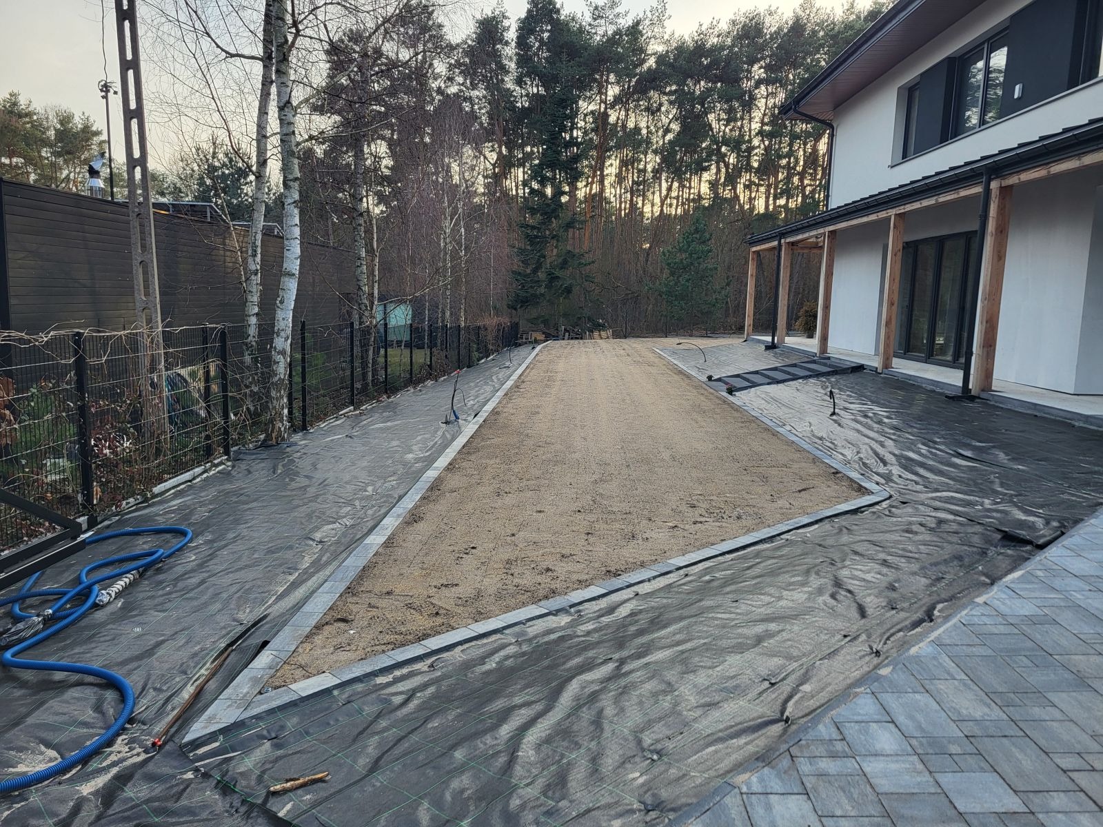 Przygotowany teren pod trawnik z rolki, obrzeża z kostki, agrowłóknina. Widok na dom i drzewa w tle. Etap prac przed rozłożeniem trawy.