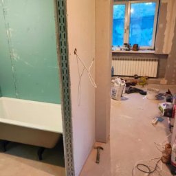 K&M BUD - Remont łazienki w trakcie: zabudowa z płyty gipsowej wokół wanny na ozdobnych nóżkach. Widoczne narzędzia, przewody i okno z grzejnikiem w tle.