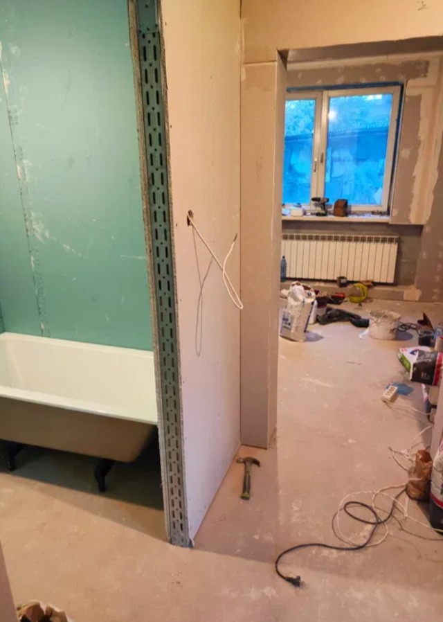 Remont łazienki w trakcie: zabudowa z płyty gipsowej wokół wanny na ozdobnych nóżkach. Widoczne narzędzia, przewody i okno z grzejnikiem w tle.