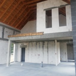 Łukasz Wykończenia - Surowy stan domu: białe bloczki, drewniany strop, betonowe elementy konstrukcyjne, widoczne instalacje. Wnętrze w trakcie tynkowania, surowy stan deweloperski.