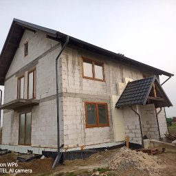 Łukasz Wykończenia - Surowy stan dwupiętrowego domu z pustaków, z widoczną izolacją, drewnianymi oknami i elementami dachu. Na pierwszym planie ziemia i palety z materiałami budowlanymi.