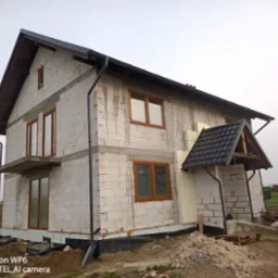 Surowy stan dwupiętrowego domu z pustaków, z widoczną izolacją, drewnianymi oknami i elementami dachu. Na pierwszym planie ziemia i palety z materiałami budowlanymi.