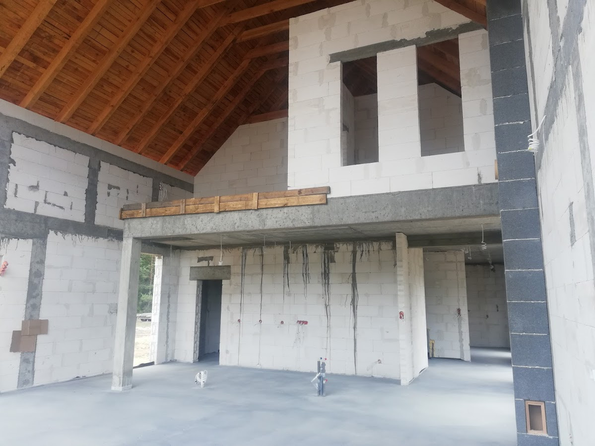 Surowy stan domu: białe bloczki, drewniany strop, betonowe elementy konstrukcyjne, widoczne instalacje. Wnętrze w trakcie tynkowania, surowy stan deweloperski.