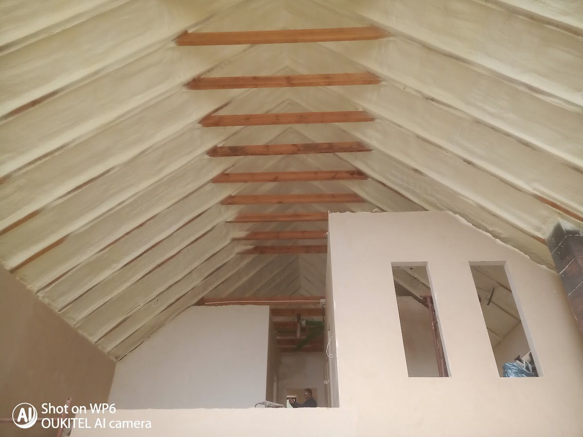 Poddasze z drewnianą konstrukcją dachu pokryte jasną pianą izolacyjną, widoczne fragmenty ścian. Ujęcie z dołu podkreśla strukturę i detale izolacji.