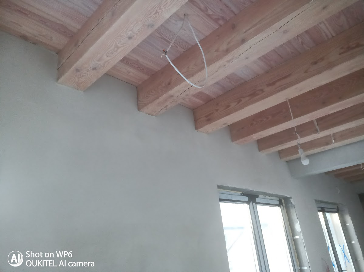 Ściana po tynkowaniu z widocznymi drewnianymi belkami stropowymi i oknami. Surowy stan wykończenia wnętrza, widoczne kable i żarówka.