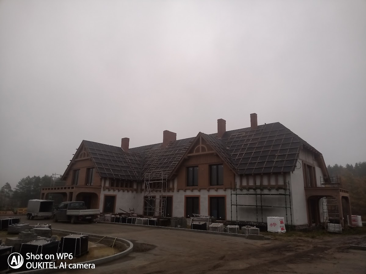 Hotel w trakcie wykończenia: widoczne rusztowania, dach z elementami deskowania, brakujące fragmenty tynku. Pochmurna pogoda, elementy krajobrazu.