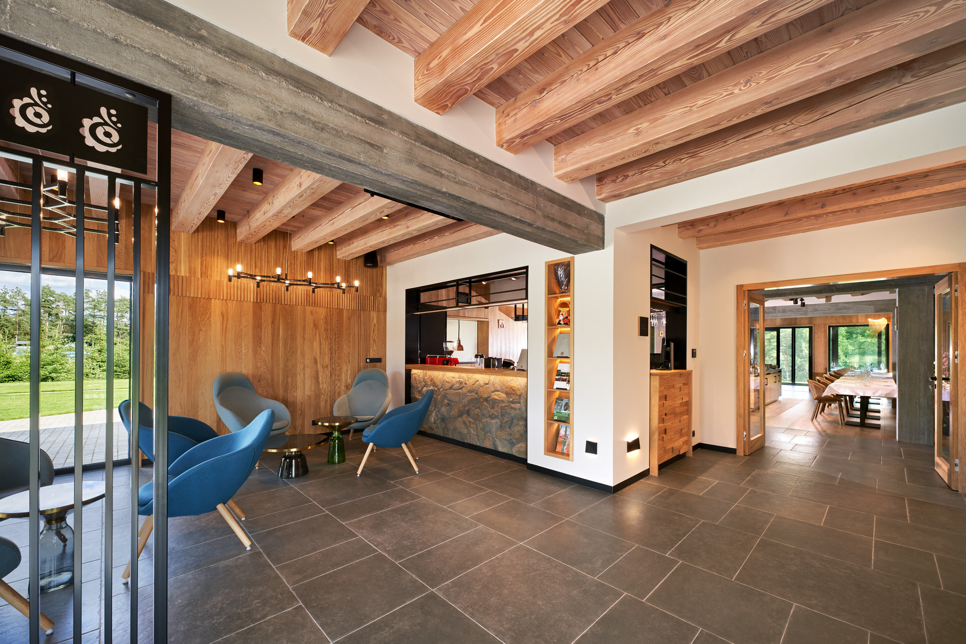 Nowoczesne wykończenie wnętrza hotelu z drewnianym sufitem, stylowymi krzesłami i widokiem na zieleń. Charakterystyczne oświetlenie i kamienna okładzina baru.