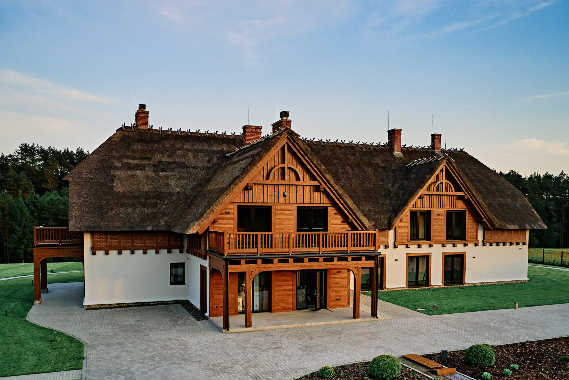 Hotel w stylu rustykalnym z drewnianą elewacją i strzechą, otoczony zielenią. Widoczny dziedziniec z kostki brukowej i zadbany trawnik. Ujęcie z lotu ptaka.