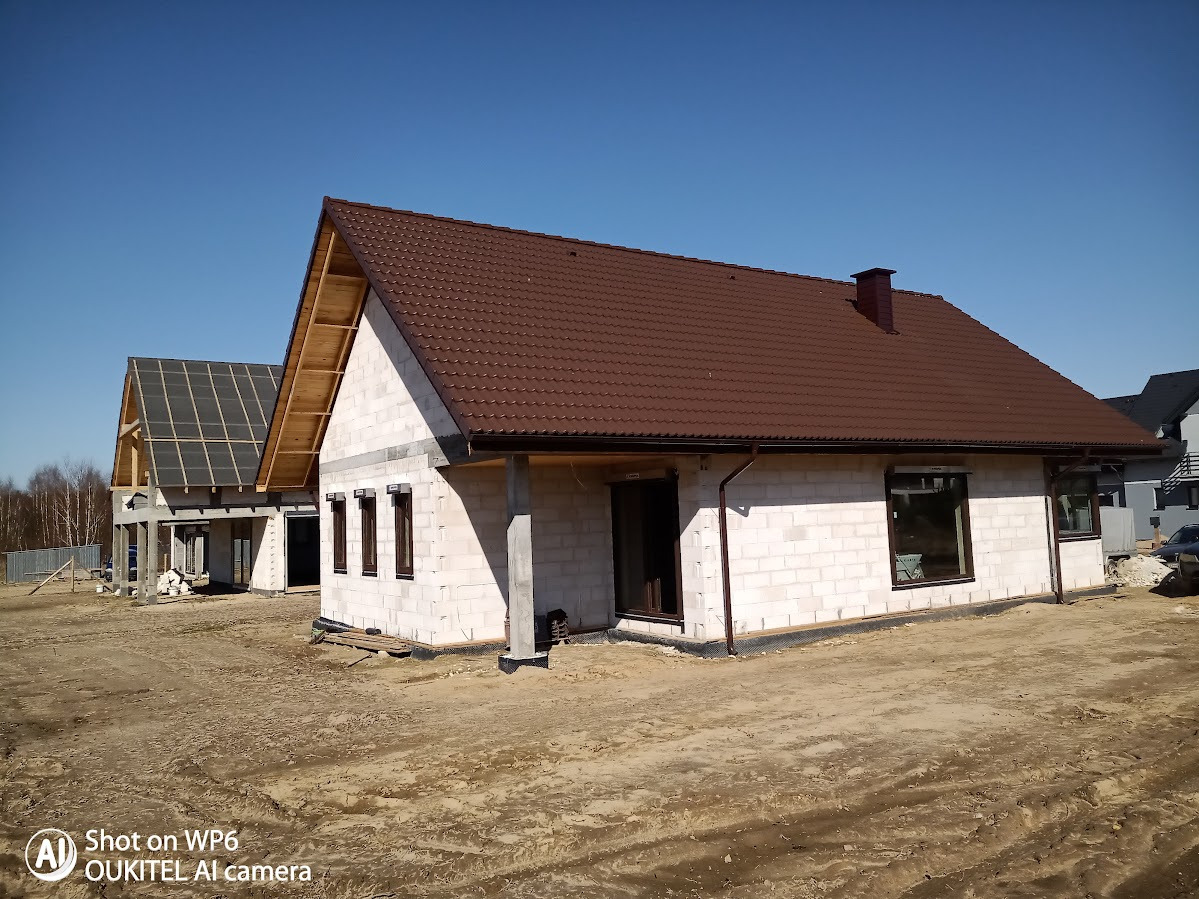 Surowy budynek jednorodzinny z brązowym dachem w trakcie budowy, widoczne bloczki betonowe i drewniana konstrukcja dachu na tle błękitnego nieba.