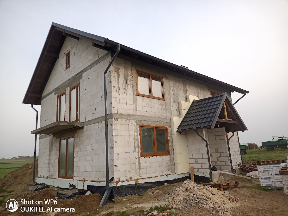 Surowy stan dwupiętrowego domu z pustaków, z widoczną izolacją, drewnianymi oknami i elementami dachu. Na pierwszym planie ziemia i palety z materiałami budowlanymi.