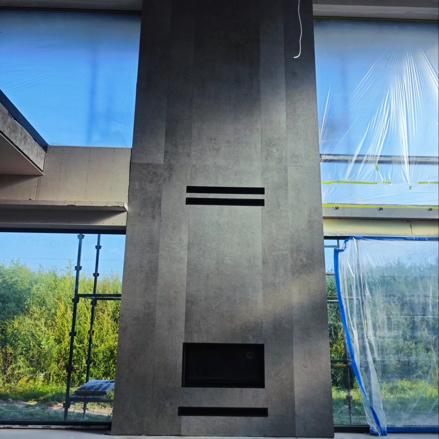 Nowoczesny kominek obłożony szarą płytą, w trakcie montażu. Okna zabezpieczone folią malarską. Widoczne rusztowanie za oknem.