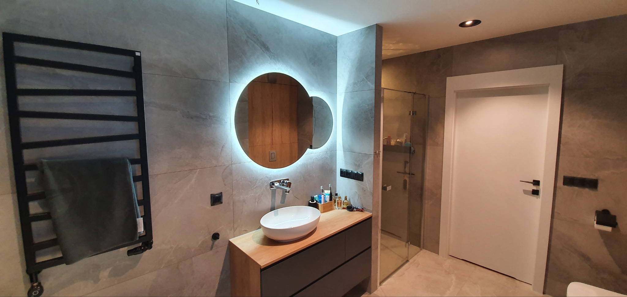 Nowoczesna łazienka z okrągłym lustrem podświetlanym LED, umywalką na szafce z drewnianym blatem i grzejnikiem drabinkowym. Szare płytki imitujące marmur.