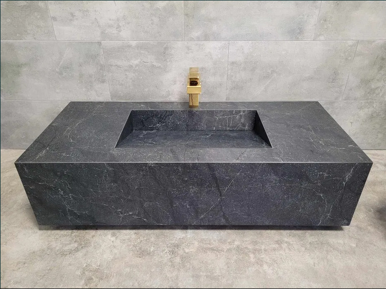 Nowoczesna, prostokątna umywalka z ciemnego kamienia z złotą baterią na tle szarych płytek. Minimalistyczny design łazienki.