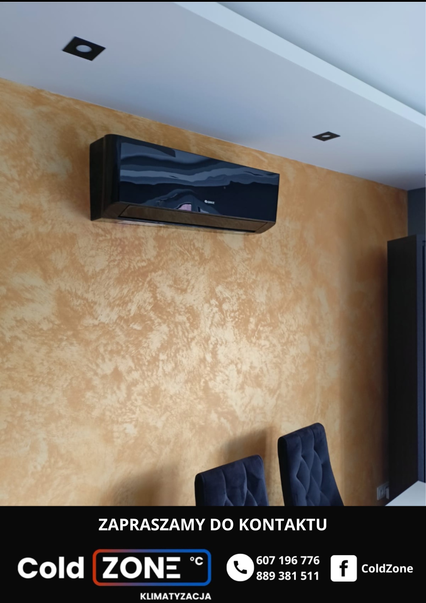 Czarna klimatyzacja Gree na ścianie o fakturze trawertynu, z widocznymi dwoma krzesłami. Oświetlenie sufitowe punktowe. Zaprasza do kontaktu Cold Zone.