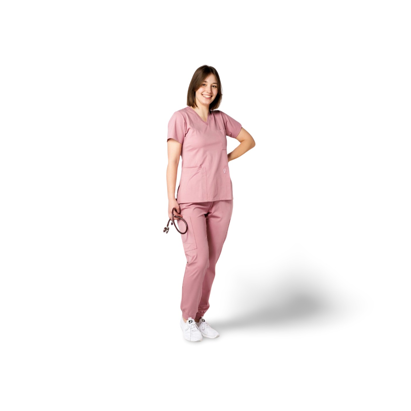 Uśmiechnięta kobieta w różowym komplecie medycznym trzyma stetoskop. Całość na białym tle, widoczne białe buty i cień. Profesjonalny strój medyczny.